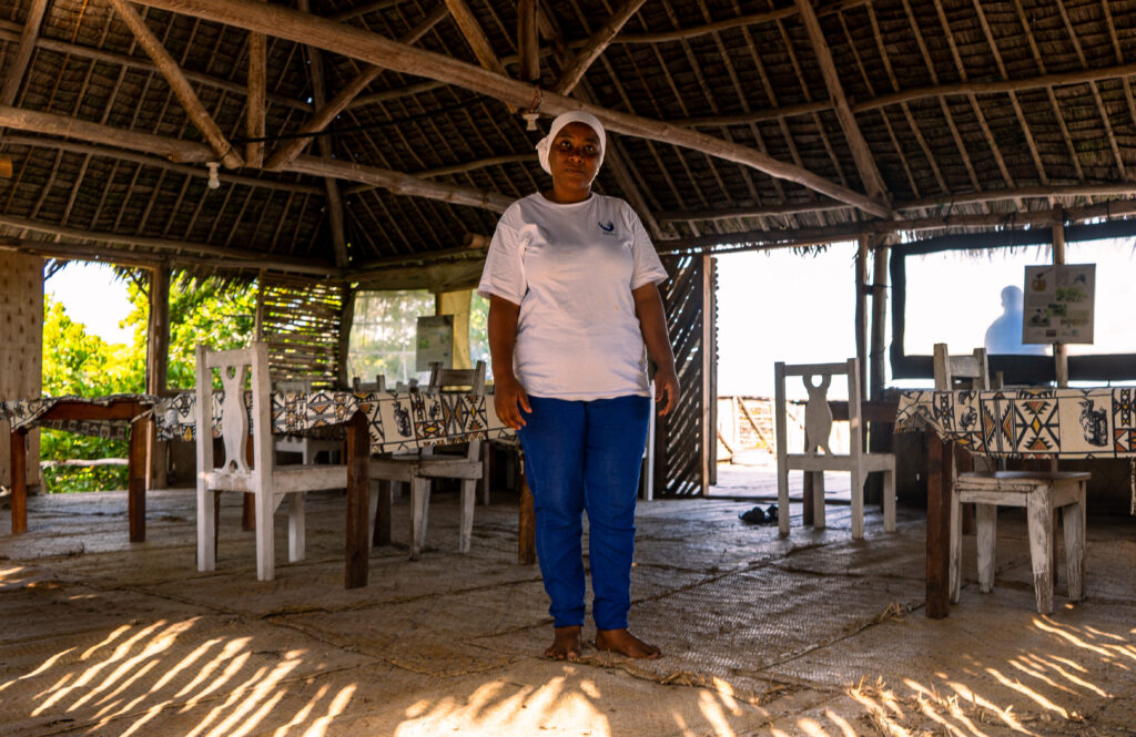 Salma Abdalla staat midden in haar restaurant. De zon schijnt naar binnen. Salma is een zwarte vrouw en draagt een wit shirt, witte hoofddoek en blauwe spijkerbroek. 