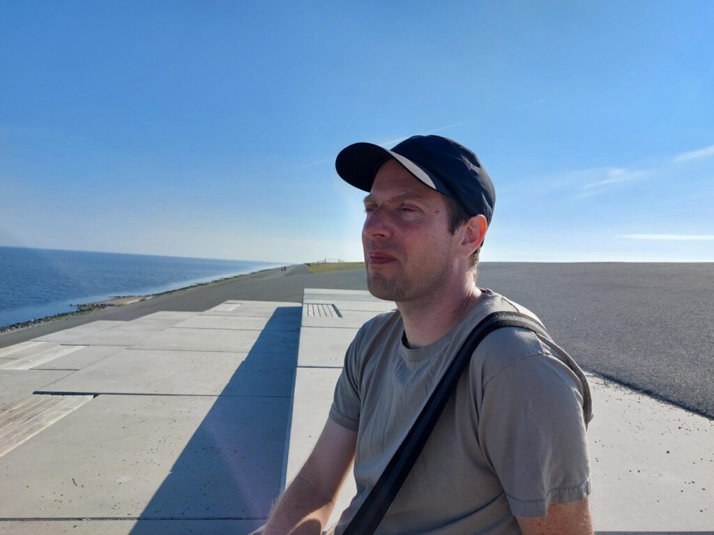 Machiel van Veen kijkt vanaf een pier uit over zee. Hij draagt een zwart petje, grijs shirt en een zwarte schoudertas.